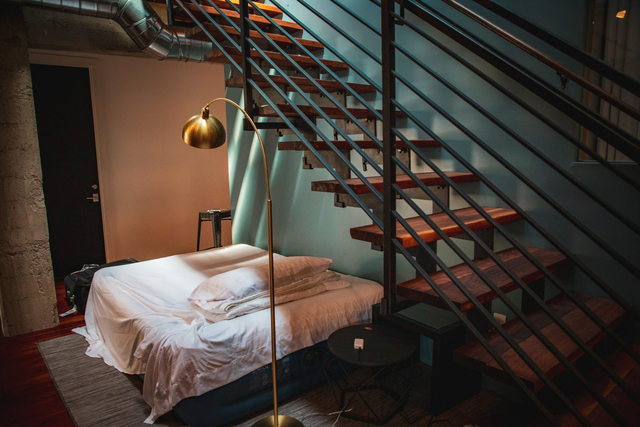 The image reveals a loft-style room with a bed under wooden stairs, a gold floor lamp, and a compact black table. The background includes exposed pipes and a door.